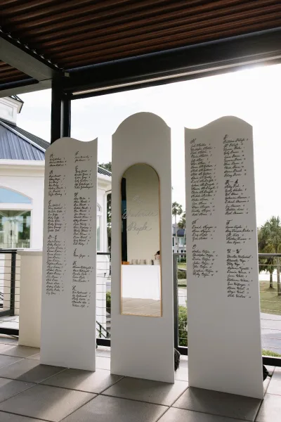 Wedding seating chart mirror with calligraphy lettering on a gold-framed arched mirror, displayed on a covered patio with palm trees beyond