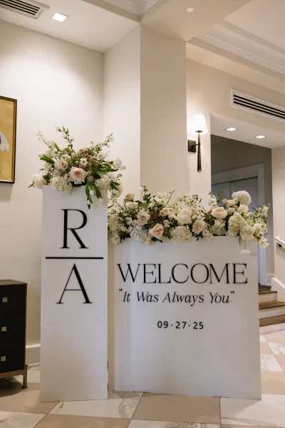 Wedding welcome sign with roses and hydrangeas greenery and monogram letters, displayed in an elegant indoor lobby with staircase