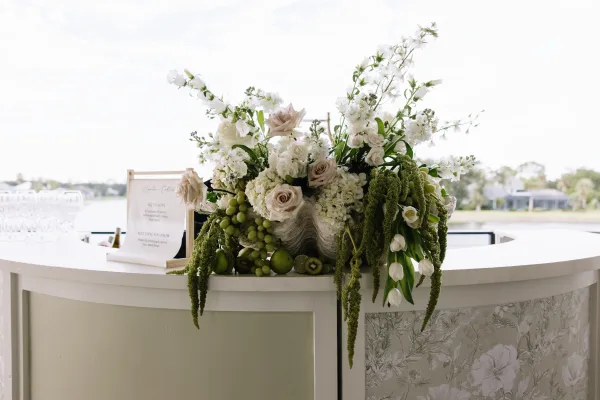 Wedding bar decor with cocktail bar floral arrangement of white roses, greenery, amaranthus, grapes, and oyster shells by a waterfront backdrop