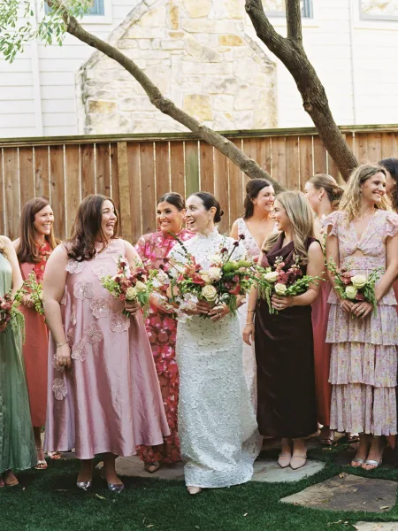 Bride and bridesmaids in mismatched bridesmaid dresses smile with blush and burgundy bouquets by a wooden fence and garden tree on a sunny lawn