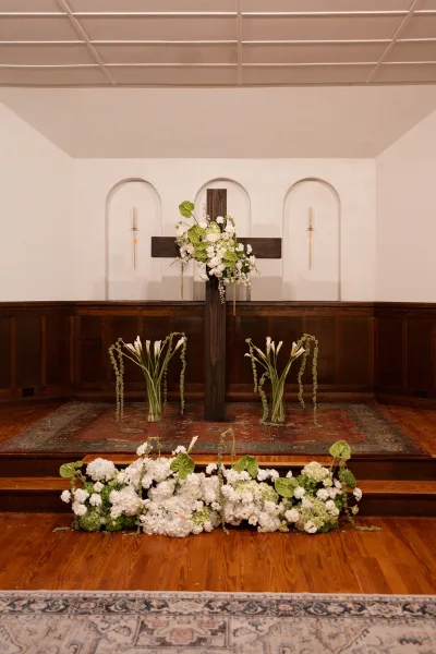 Ceremony altar decor featuring a church cross floral arrangement with a row of candles, set before arched chapel niches and wood paneling.
