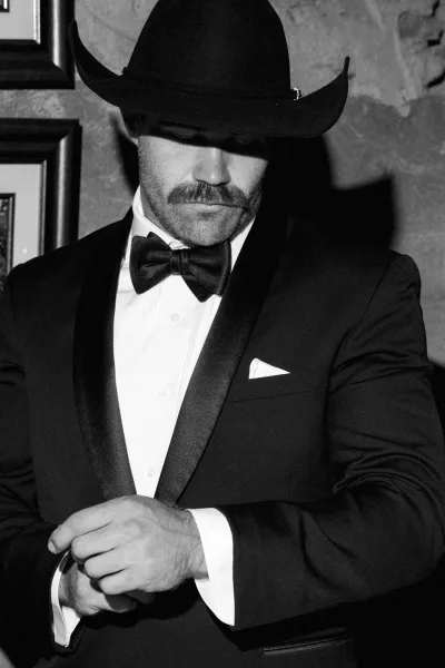 Groom portrait of a tuxedo groom posed against a wall with framed artwork, calm expression in soft, flattering light for a timeless feel