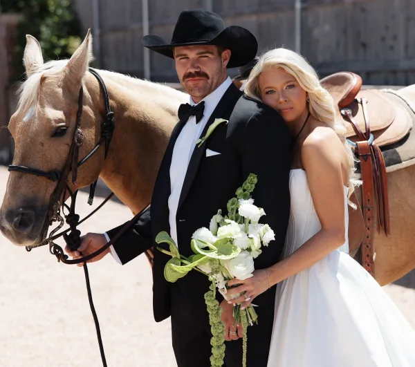 Couple portrait of a bride leaning on her groom in a cowboy hat, posed by a rustic barn wall and wooden fence with gravel and greenery.
