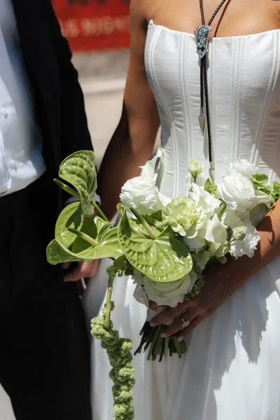 Bridal bouquet in green and white with anthurium leaves, held by the bride against a red urban wall on a concrete urban street.