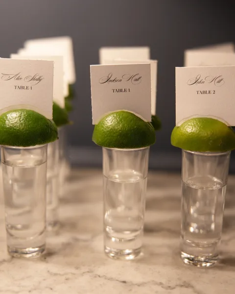 Escort card display of shot glass escort cards arranged with limes on a marble countertop against a gray wall.