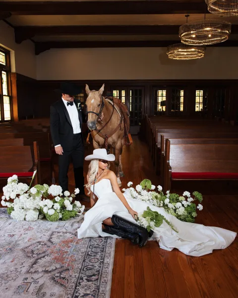 Couple portrait with a bride in cowboy boots, standing by chapel pews beneath stained glass windows and wooden beams.