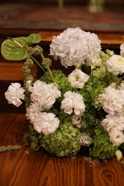 Wedding floral arrangement of white and green wedding flowers featuring green hydrangea, resting on a wood floor beside a woven rug.