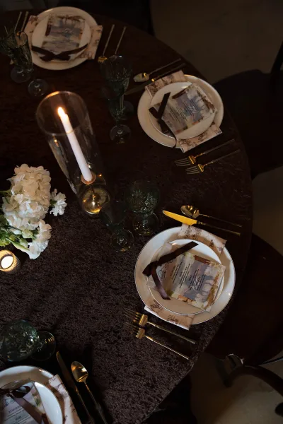 Reception tablescape showcasing an elegant wedding place setting with green goblets on a round table draped in dark linen and a wood chair nearby.