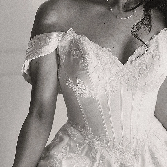 Wedding dress with off the shoulder straps and corset bodice, lace applique detailing, and a delicate necklace against a plain wall