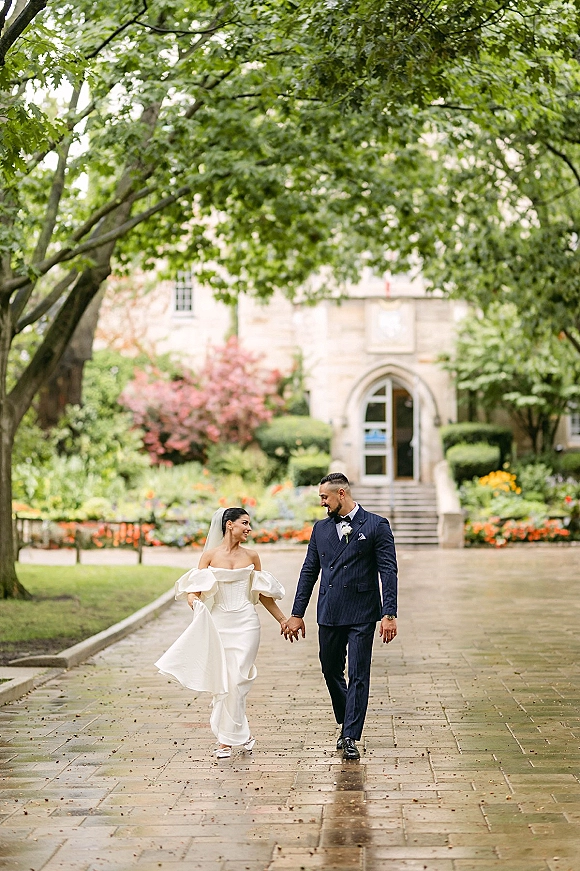 Couple portrait of bride in off-the-shoulder wedding dress and groom in suit holding hands on a stone garden walkway under trees