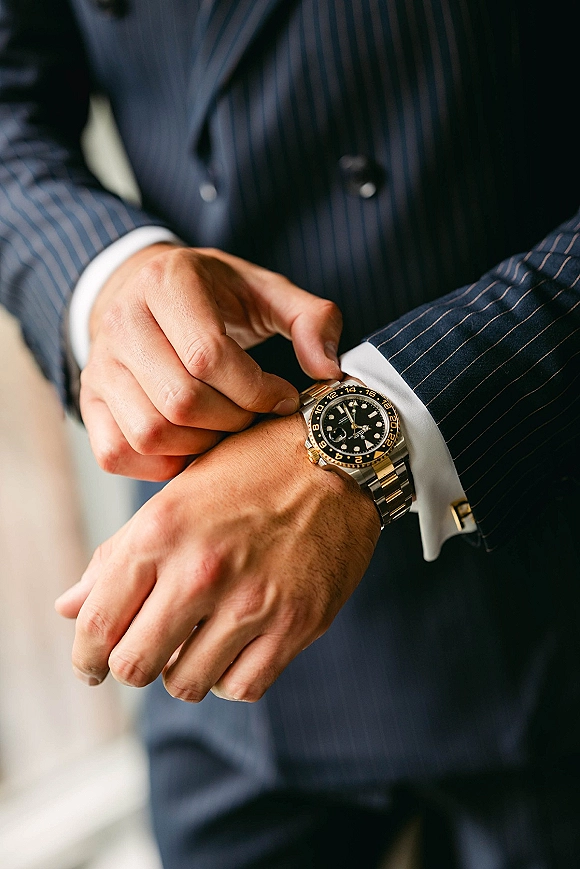 Groom watch on his wrist as he adjusts a gold and silver wristwatch over a white shirt cuff, pinstripe suit and cufflinks in soft indoor light