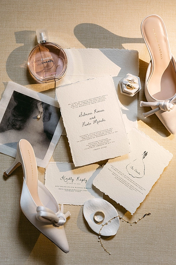 Invitation suite with deckle edge wedding invitations, RSVP and details cards, rings, pearl jewelry, perfume, and heels on linen in sun shadows