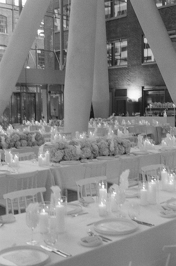 Reception tablescape with white linens, taper candles and glass votives, low floral centerpieces and place settings in an industrial loft with brick walls