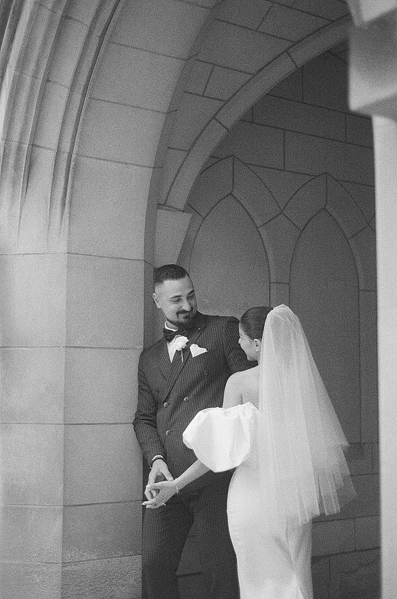 Couple portrait in a black and white wedding portrait, bride and groom holding hands under a stone archway with a long veil