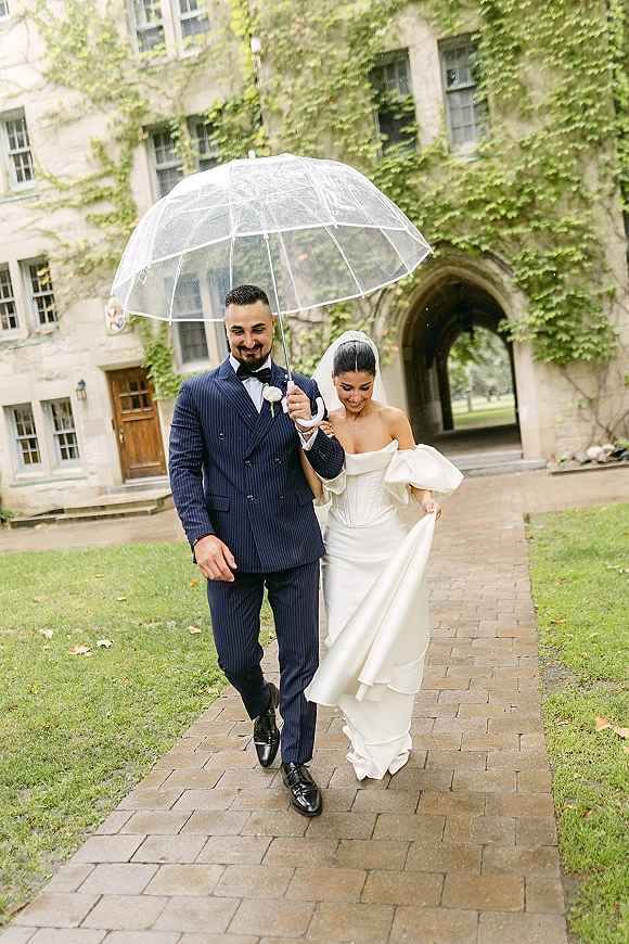 Couple portrait of bride and groom under a clear umbrella, her veil flowing, strolling by an ivy-covered stone archway on brick path