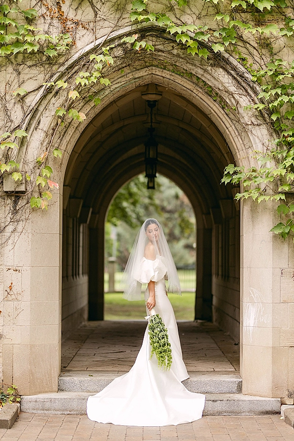 Bridal portrait of a cathedral veil bride holding a cascading greenery bouquet beneath an ivy-draped stone archway with lanterns
