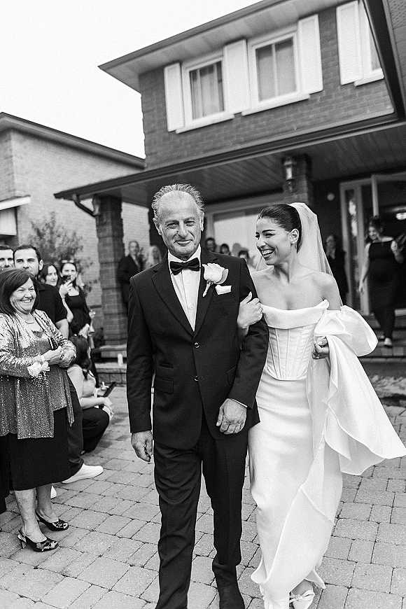 Processional moment as bride walks arm in arm with her father in a strapless dress and long veil past guests outside a brick house porch