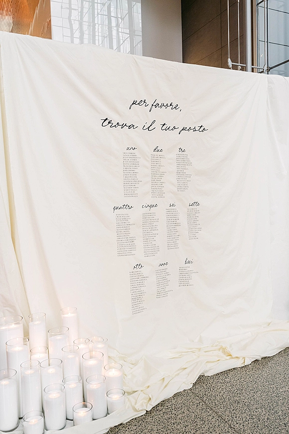Seating chart sign with wedding seating chart calligraphy, draped fabric backdrop, and pillar candles in glass hurricanes by large windows