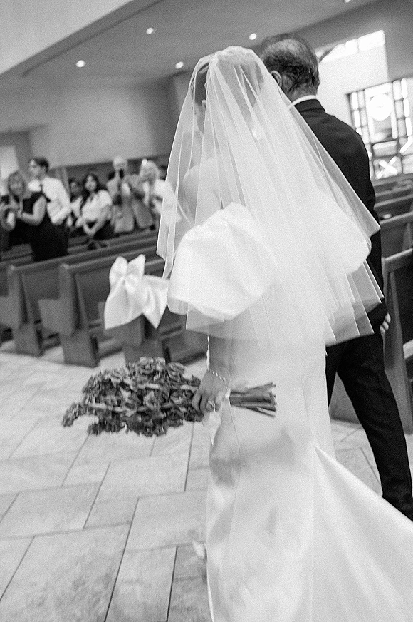 Wedding processional as bride walking down aisle with bouquet and long veil, escorted by her father past pew bows in a church aisle