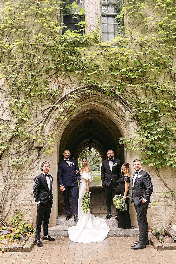 Wedding party portrait with bride holding cascading greenery bouquet, bridesmaids and groomsmen posed by an ivy-covered stone archway