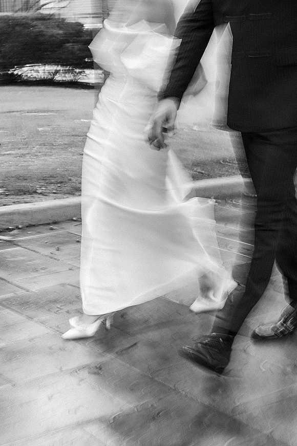 Wedding couple walking hand in hand away on a city sidewalk, bride’s veil and dress train flowing beside groom in a dark suit