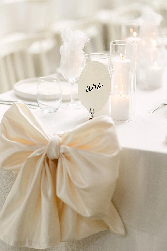 Reception tablescape with wedding table number in calligraphy beside pillar candles in glass hurricanes on a white tablecloth with place settings
