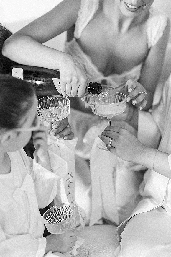 Bridal champagne toast with champagne bottle pouring into coupe glasses, bride’s manicured hands showing wedding ring over bed linens indoors