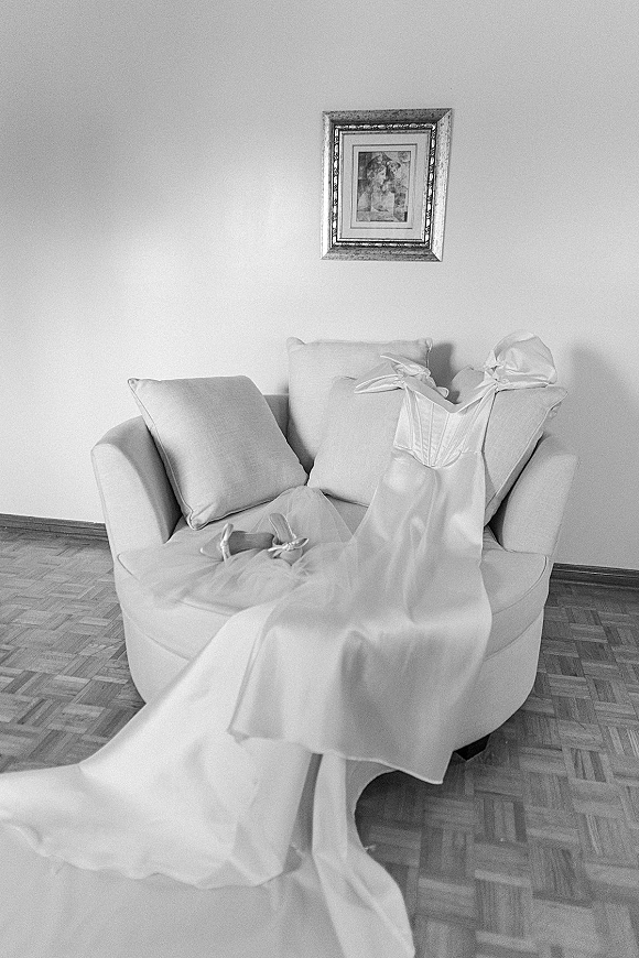 Wedding dress, strapless satin gown with corset bodice and long train draped on a sofa with tulle veil and bridal shoes on parquet floor