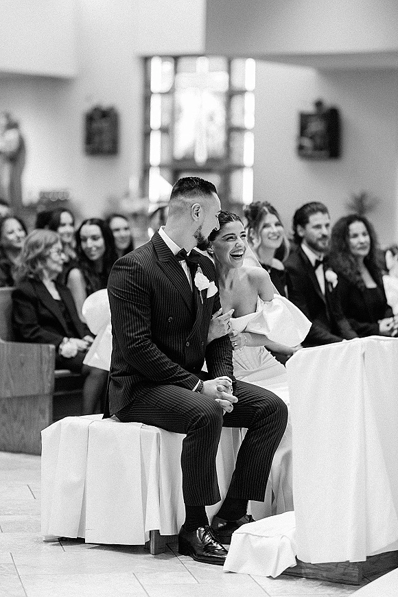 Ceremony moment as bride laughs in her veil holding the groom’s arm while seated in a chapel, guests watching from benches in window light