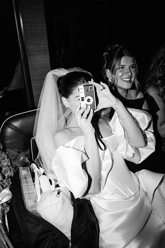 Bride candid moment with film camera, veil draped over an off-the-shoulder gown, seated on a leather couch in a dark wood-paneled room