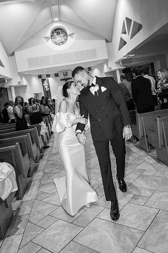 Wedding recessional as bride and groom walk hand in hand down a church aisle, smiling past guests, with pew bows and stained glass behind