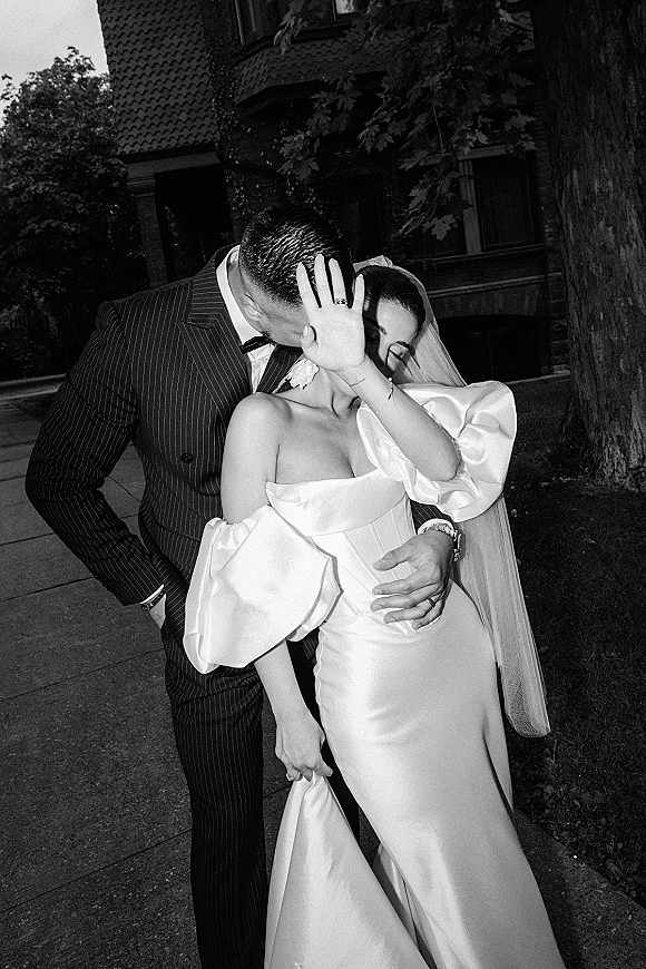 Wedding kiss portrait of bride and groom kissing, her veil and off-shoulder satin dress beside a stone building and trees outdoors