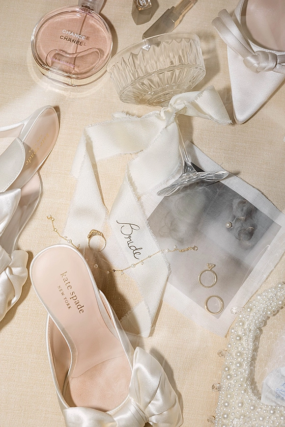 Bridal details flatlay with a wedding ring flatlay, pearl jewelry, satin ribbon, perfume, makeup, and shoes arranged on linen fabric