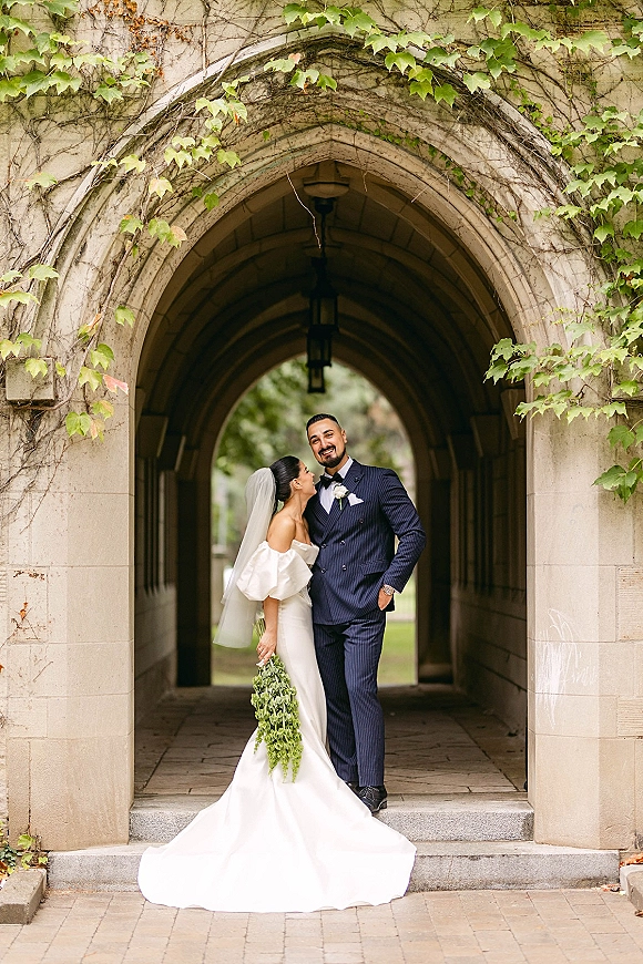 Couple portrait of bride in off-shoulder gown and veil with cascading greenery bouquet, leaning on groom under ivy stone archway lanterns