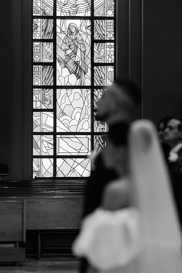 Wedding kiss portrait of bride and groom kissing in a church, veil draped over his suit jacket with stained glass light and wooden pews behind