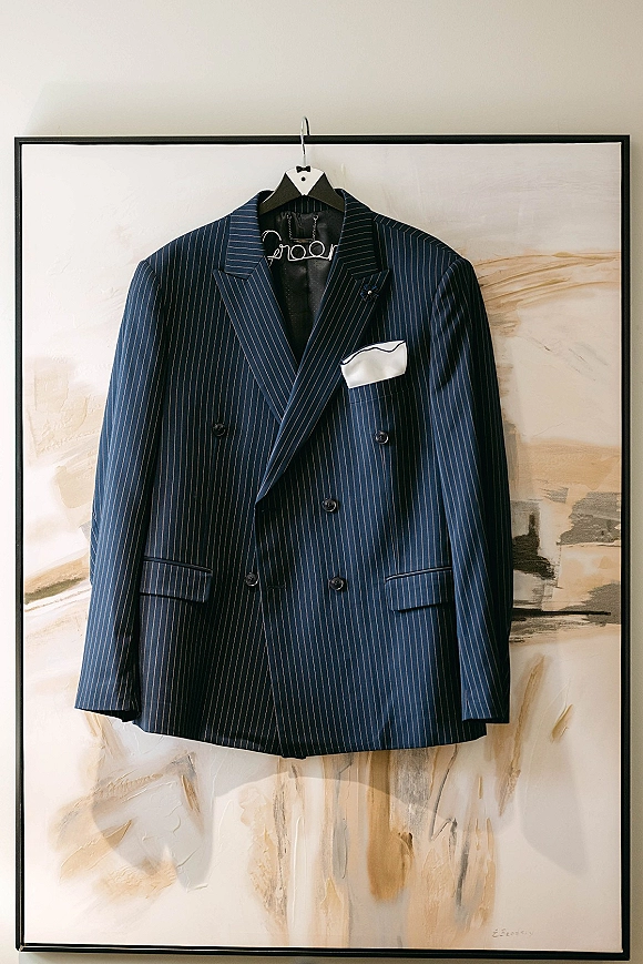 Groom suit in a navy pinstripe suit jacket hanging on a hanger, double breasted with a white pocket square and lapel pin by abstract art