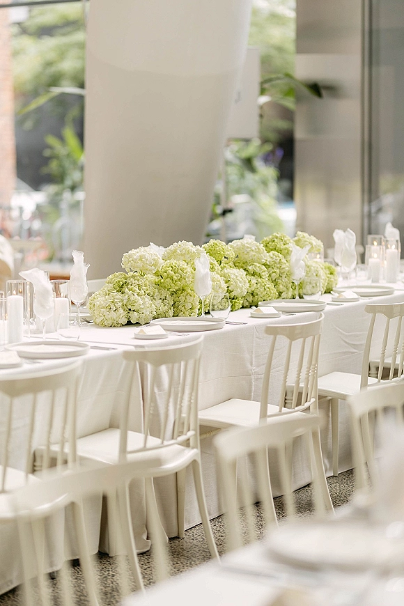 Reception tablescape on a long banquet table with green hydrangea centerpiece, cylinder and taper candles, in a modern venue by large windows