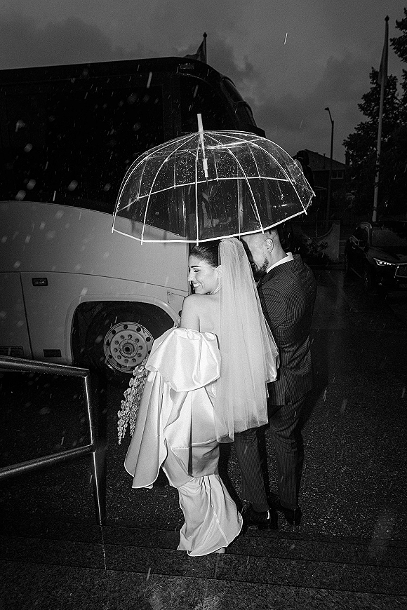 Couple portrait in a rainy wedding portrait, bride in satin dress and veil with bouquet as groom hugs her under a clear umbrella on a street