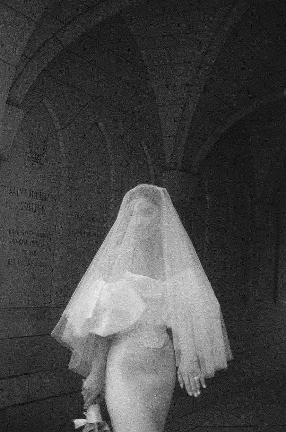 Bridal portrait in black and white of a bride in a cathedral veil, holding a bouquet under a stone archway with deep corridor shadows