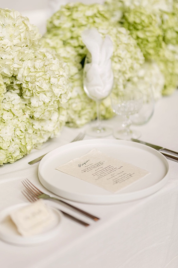 Reception tablescape with a wedding place setting featuring a menu card on a white plate, silver flatware, glassware, and hydrangeas on white linen