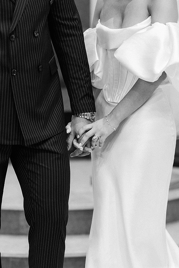 Couple holding hands, wedding hands close up showing engagement ring and band beside bride’s bracelet and groom’s pinstripe suit cuff on indoor steps