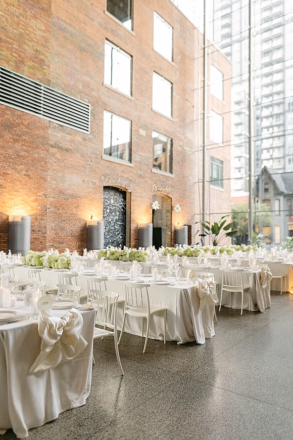 Reception tablescape with white wedding reception tables, candles, glassware, and white floral centerpieces beside brick wall and tall windows.