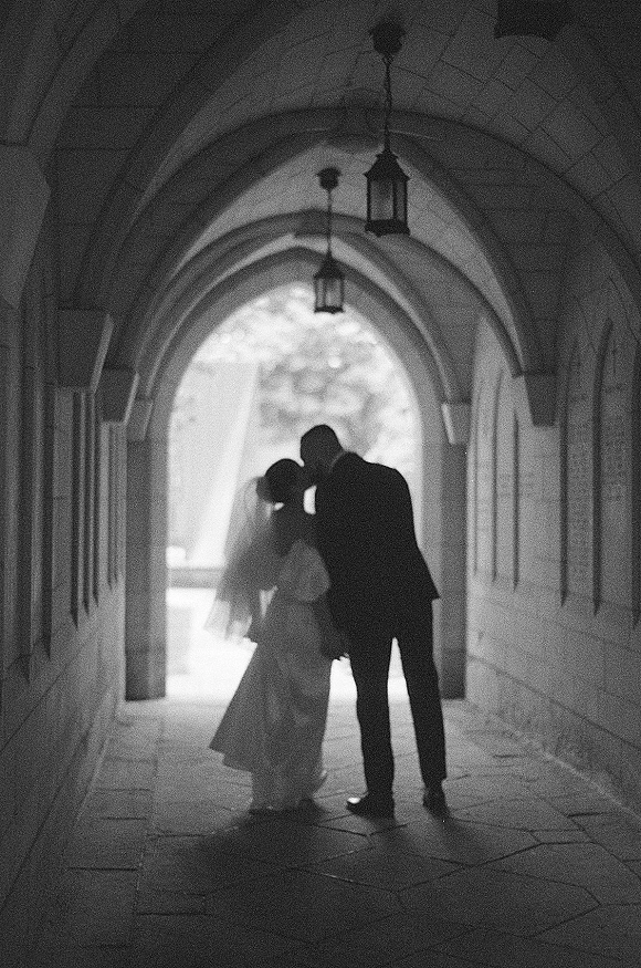 Wedding kiss silhouette of bride and groom in black and white, veil flowing as they stand beneath a stone archway with lanterns and backlit trees