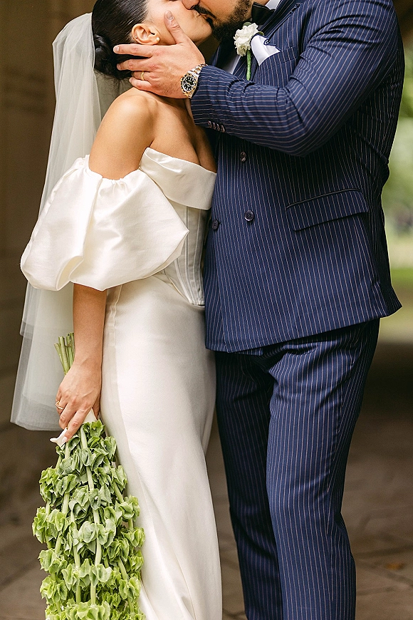 Wedding kiss portrait of bride and groom kiss as he cradles her face, bridal veil flowing, outdoors on a stone walkway with greenery