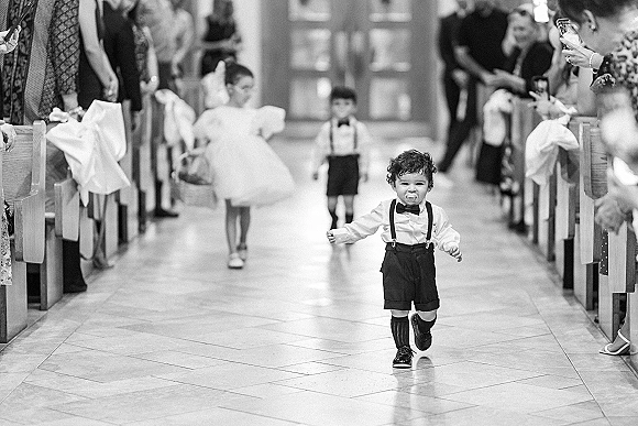 Wedding processional with a ring bearer walking down aisle in suspenders and bow tie, flower girl beside him in a church aisle with pew bows
