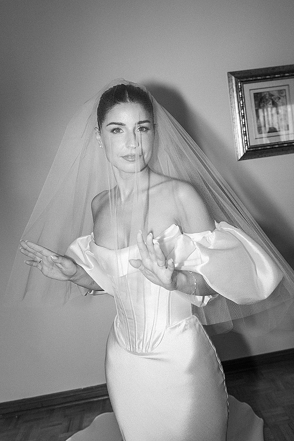 Bridal portrait in black and white of a bride with veil over her face, wearing an off-shoulder satin corset gown indoors by framed artwork
