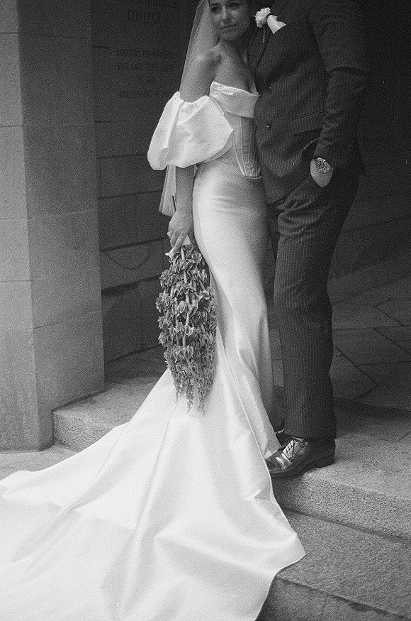Couple portrait in black and white, bride leaning on groom with veil over face and cascading bouquet on stone steps by wall