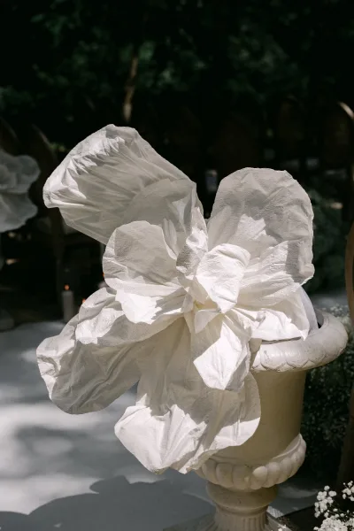 Ceremony aisle decor with a white fabric flower on an urn pedestal and candle, lining a sunlit outdoor walkway with green foliage