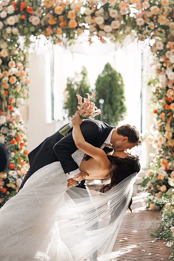 Wedding kiss portrait of groom dipping bride in strapless lace dress and veil, holding calla lily bouquet under floral arch on brick aisle