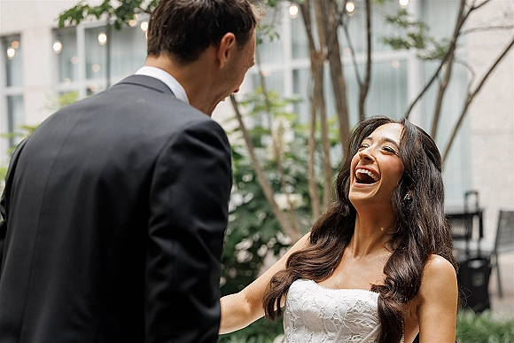 Wedding couple moment as the bride laughs candid, looking up at the groom in a courtyard with string lights and greenery behind them